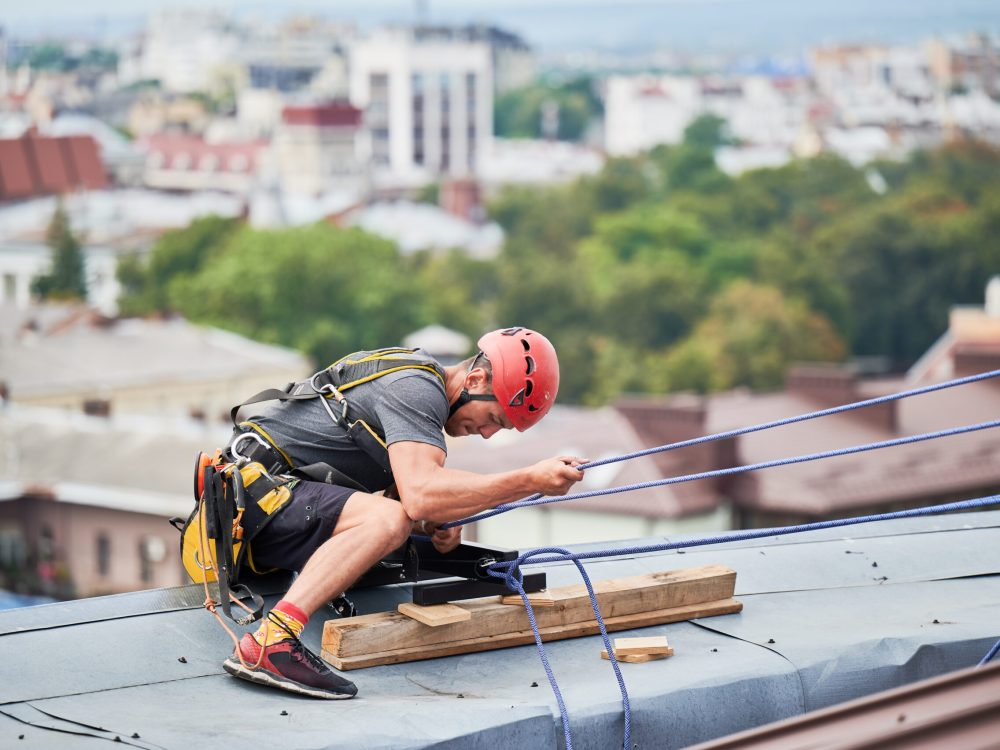 Industrial mountaineering worker in protective helmet using safety climbing rope while climbing building roof. Man climber using safety climbing equipment during industrial high-rise work.
